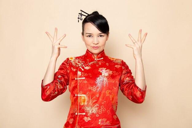 Una vista frontal hermosa geisha japonesa en el tradicional vestido rojo japonés con palos de pelo posando con las manos de pie sobre la ceremonia de fondo crema entretenido Japón oriental