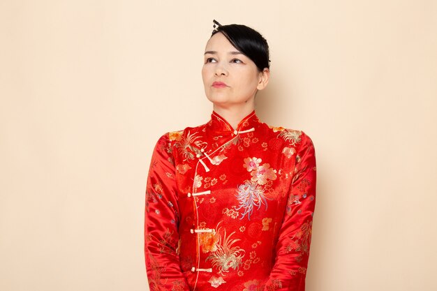 Una vista frontal hermosa geisha japonesa en el tradicional vestido rojo japonés con palos de pelo posando de pie en la ceremonia de fondo crema entretenido Japón