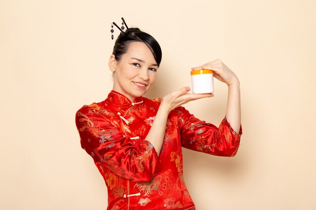 Una vista frontal hermosa geisha japonesa en el tradicional vestido rojo japonés con palos de pelo posando sonriendo feliz celebración crema puede en la ceremonia de fondo crema Japón