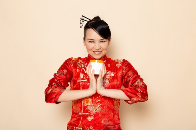 Una vista frontal hermosa geisha japonesa en el tradicional vestido rojo japonés con palos de pelo posando sonriendo feliz celebración crema puede en la ceremonia de fondo crema Japón