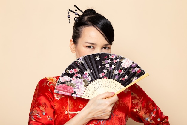 Foto gratuita una vista frontal hermosa geisha japonesa en tradicional vestido rojo japonés con palos de pelo posando sosteniendo abanico plegable elegante en la ceremonia de fondo crema japón