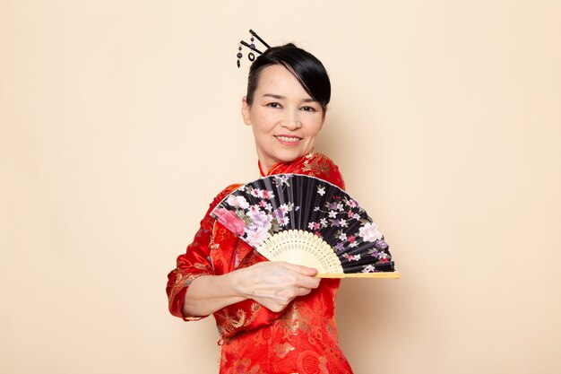 Una vista frontal hermosa geisha japonesa en tradicional vestido rojo japonés con palos de pelo posando sosteniendo abanico plegable elegante sonriendo en la ceremonia de fondo crema Japón