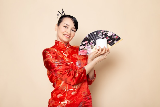 Foto gratuita una vista frontal hermosa geisha japonesa en el tradicional vestido rojo japonés con palos de pelo posando sosteniendo abanico plegable y vela blanca elegante en la ceremonia de fondo crema japón