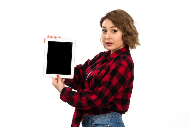 Una vista frontal hermosa joven en camisa a cuadros rojo-negro y jeans azul con tableta blanca sobre el blanco