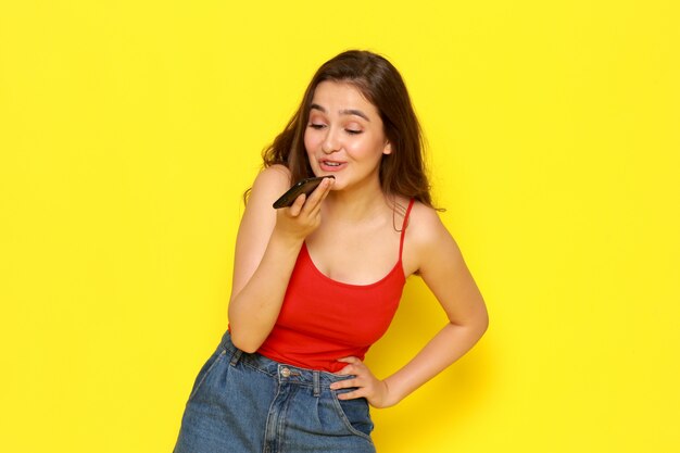 Una vista frontal hermosa joven en camisa roja y jeans azul enviando mensaje de voz