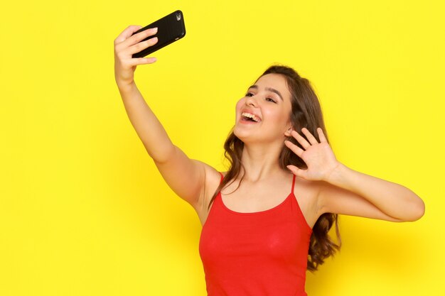 Una vista frontal hermosa joven en camisa roja y jeans azul tomando un selfie con una sonrisa