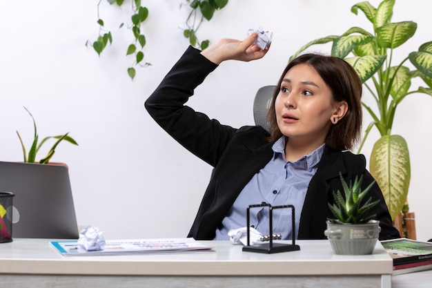 Una vista frontal hermosa joven empresaria en chaqueta negra y camisa azul jugando y lanzando bolas de papel frente a la mesa de trabajo de oficina de negocios