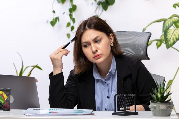 Una vista frontal hermosa joven empresaria en chaqueta negra y camisa azul pensando delante de la mesa de trabajo de oficina de negocios