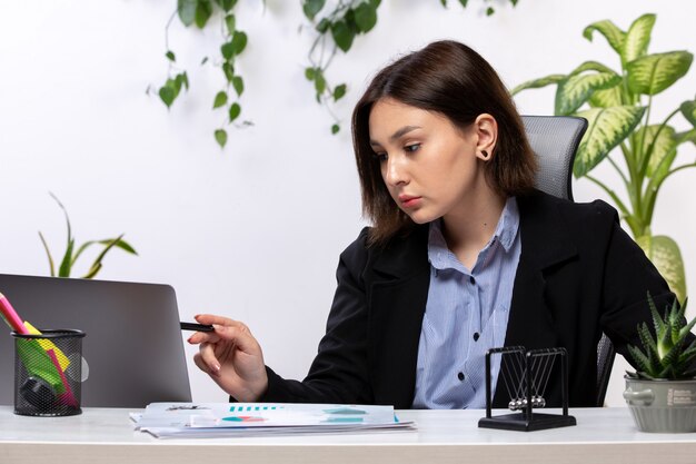 Una vista frontal hermosa joven empresaria en chaqueta negra y camisa azul que trabaja con la computadora portátil frente a la mesa de trabajo de la oficina de negocios