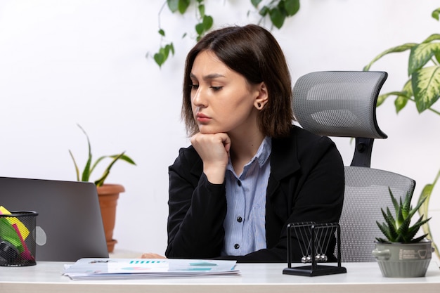 Una vista frontal hermosa joven empresaria en chaqueta negra y camisa azul sintiendo ansiedad frente a la mesa de trabajo de la oficina de negocios