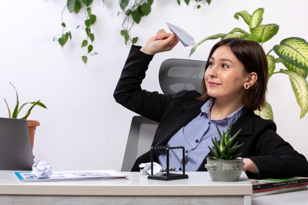 Una vista frontal hermosa joven empresaria en chaqueta negra y camisa azul sonriendo arrojando aviones de papel frente a la mesa de trabajo de la oficina de negocios