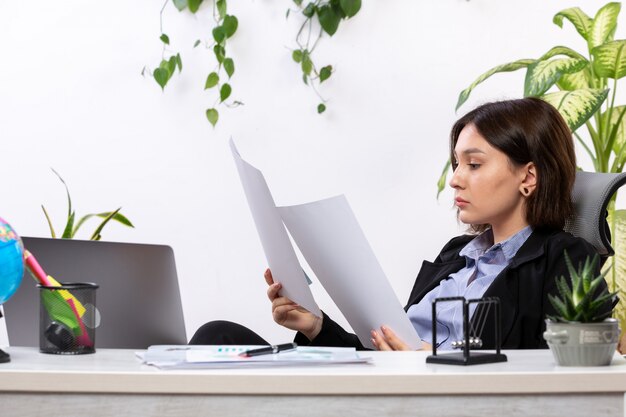 Una vista frontal hermosa joven empresaria en chaqueta negra y camisa azul trabajando con documentos frente a la mesa de trabajo de la oficina de negocios
