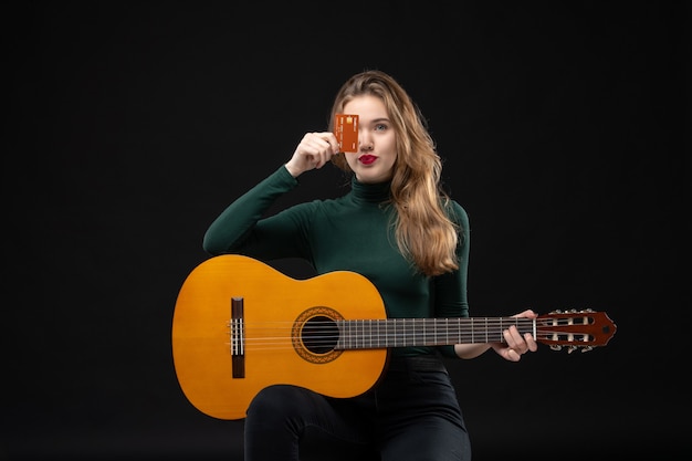 Vista frontal de la hermosa joven músico emocional divertido sosteniendo la guitarra y mostrando la tarjeta bancaria en la oscuridad