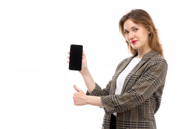 Una vista frontal hermosa jovencita en camiseta blanca jeans negros y abrigo sosteniendo mostrando smartphone sonriendo en el blanco