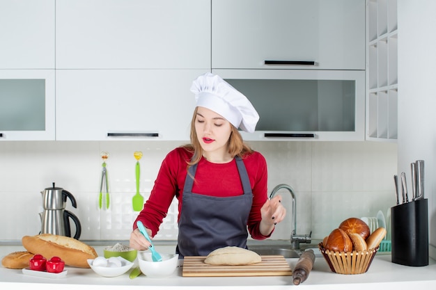 Vista frontal hermosa mujer joven con sombrero de cocinero y delantal batiendo huevos en la cocina
