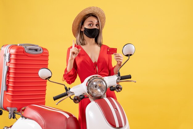 Vista frontal de la hermosa mujer con máscara negra con tarjeta de crédito cerca de ciclomotor y maleta roja