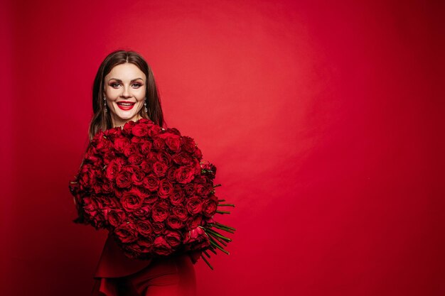Vista frontal de la hermosa mujer en vestido rojo con maquillaje manteniendo un hermoso ramo de rosas mirando a la cámara y sonriendo Bonita modelo posando sobre fondo rojo aislado Concepto de día de San Valentín