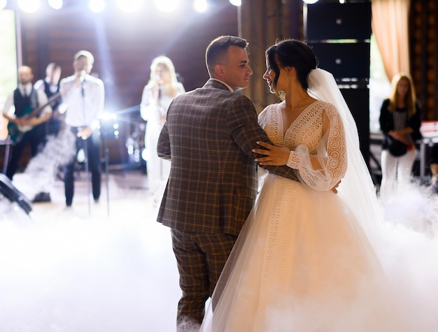 Vista frontal de la hermosa novia con un increíble vestido de novia con mangas largas sosteniendo las manos del novio y mirándolo durante el primer baile de bodas