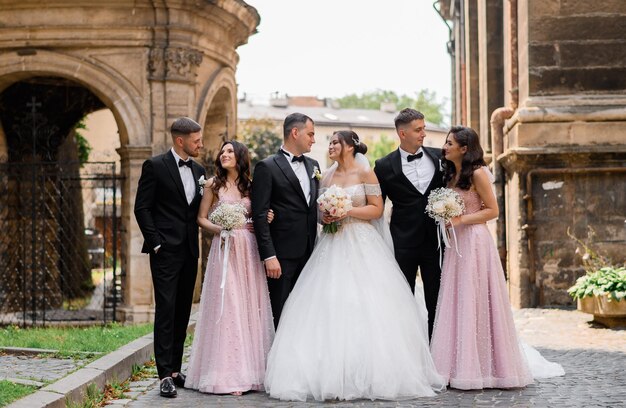 Vista frontal de la hermosa novia y el novio en ropa de boda sonriendo y mirándose mientras están de pie entre amigos