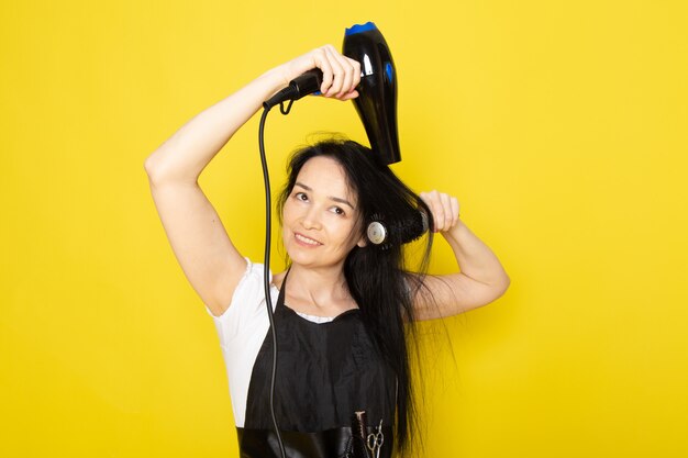 Una vista frontal hermosa peluquera en camiseta blanca capa negra con pinceles con cabello lavado secado cepillando su cabello posando y sonriendo sobre el fondo amarillo estilista peluquero