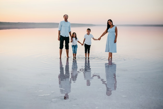 Vista frontal de hermosos padres jóvenes, su linda hijita e hijo tomados de la mano, caminando por la playa