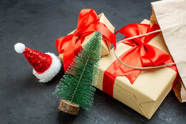 Vista frontal de hermosos regalos con cinta roja y árbol de navidad sombrero de santa claus en una mesa oscura