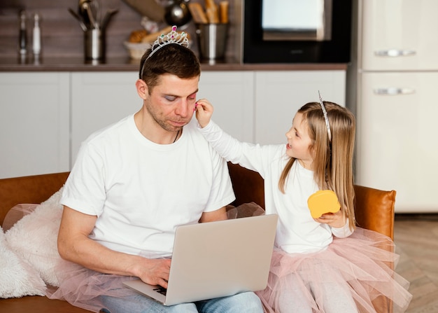 Vista frontal de la hija jugando con el padre mientras trabaja en la computadora portátil