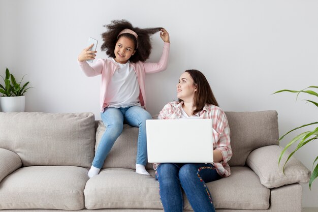 Vista frontal hija tomando una foto mientras la madre está trabajando