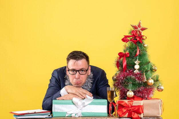 Vista frontal del hombre sin alegría sentado en la mesa cerca del árbol de Navidad y presenta en amarillo