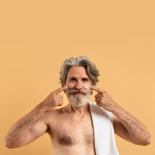 Vista frontal del hombre anciano sonriente con barba aplicando crema en la cara con espacio de copia