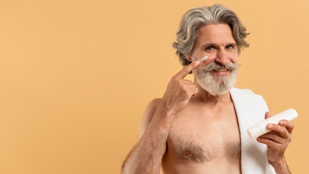 Vista frontal del hombre anciano sonriente con barba aplicando crema en la cara