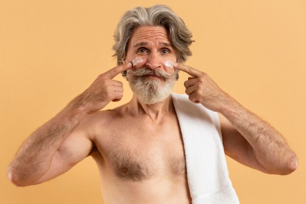 Vista frontal del hombre anciano sonriente con barba aplicando crema en la cara