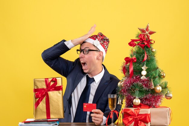 Vista frontal del hombre asombrado poniendo la mano en la frente sentado en la mesa cerca del árbol de Navidad y presenta en la pared amarilla