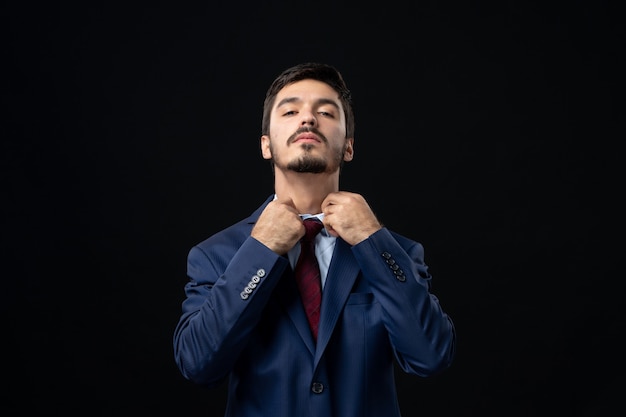 Vista frontal del hombre barbudo en traje enderezando su corbata y posando en la pared oscura