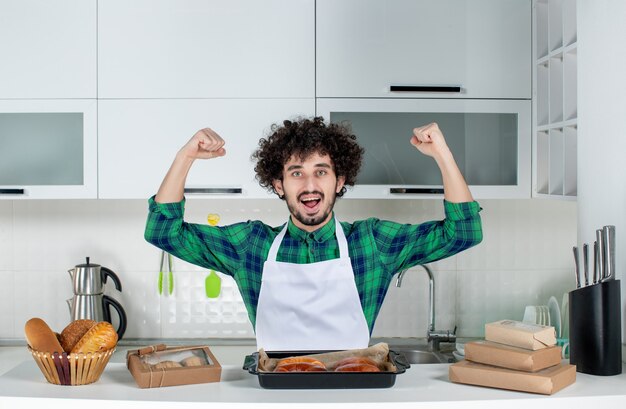 Vista frontal del hombre confiado de pie detrás de la mesa con pasteles recién horneados y mostrando su musculoso en la cocina blanca