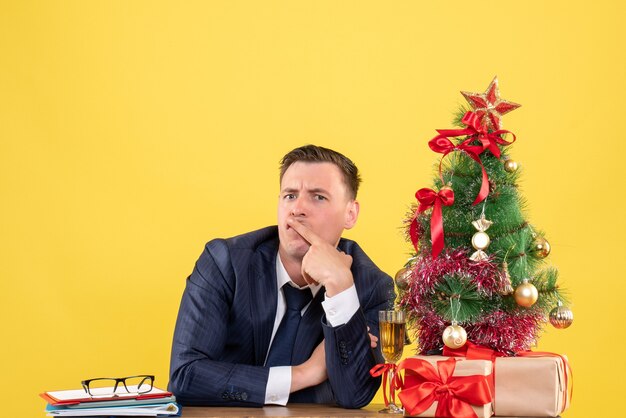 Vista frontal del hombre confundido poniendo el dedo en la boca sentado en la mesa cerca del árbol de Navidad y presenta en amarillo