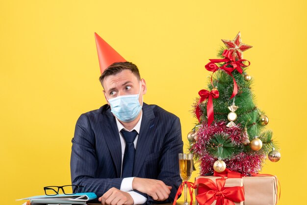 Vista frontal del hombre confundido sentado en la mesa cerca del árbol de Navidad y presenta en amarillo