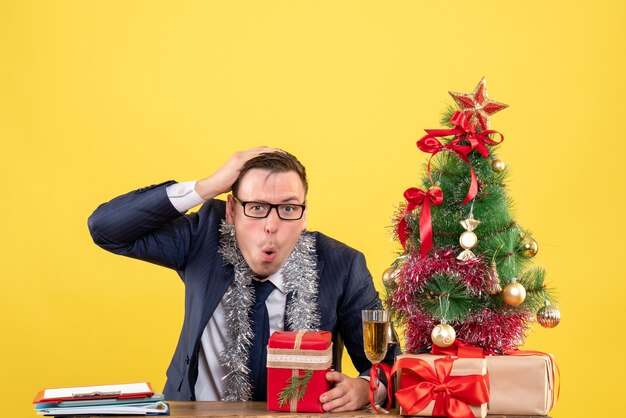 Vista frontal del hombre confundido sosteniendo su cabeza sentado en la mesa cerca del árbol de Navidad y presenta en amarillo