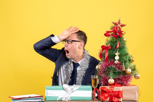 Foto gratuita vista frontal del hombre decepcionado poniendo la mano en la frente sentado en la mesa cerca del árbol de navidad y presenta en amarillo.