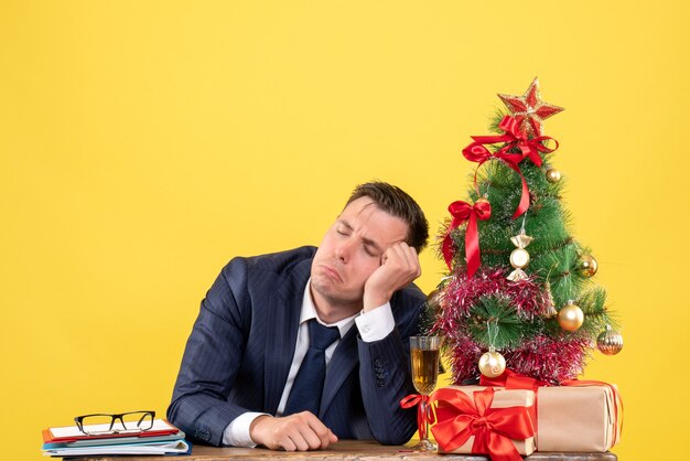 Vista frontal del hombre decepcionado sentado en la mesa cerca del árbol de Navidad y presenta en amarillo.