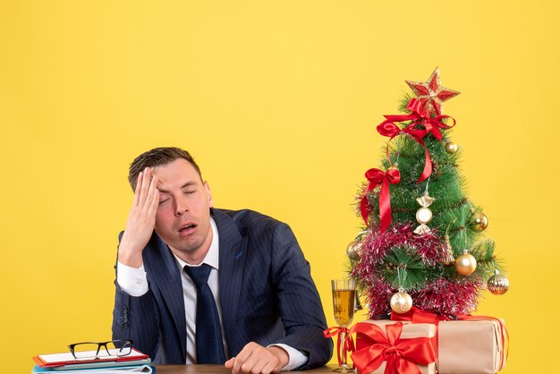 Vista frontal del hombre decepcionado sosteniendo su cabeza sentado en la mesa cerca del árbol de Navidad y regalos en amarillo.