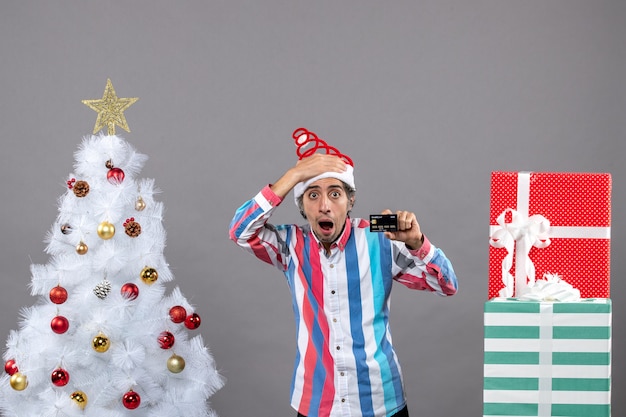 Vista frontal del hombre decepcionado con tarjeta de crédito de pie cerca del árbol de Navidad blanco