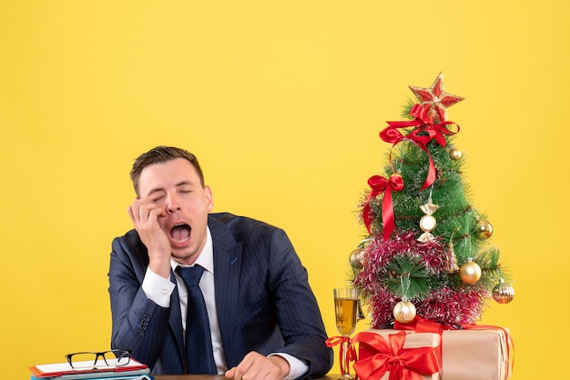 Vista frontal del hombre deprimido sentado en la mesa cerca del árbol de Navidad y regalos en amarillo.