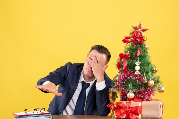 Vista frontal del hombre deprimido sentado en la mesa tapándose los ojos con la mano cerca del árbol de Navidad y regalos en amarillo