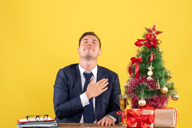 Vista frontal del hombre dichoso poniendo la mano sobre su pecho sentado en la mesa cerca del árbol de Navidad y presenta en la pared amarilla