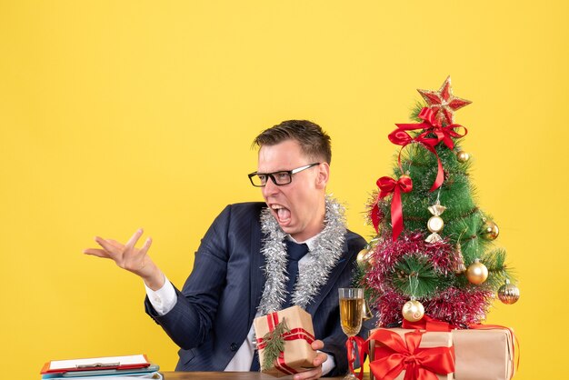 Vista frontal del hombre enojado con anteojos sentado en la mesa cerca del árbol de Navidad y presenta en la pared amarilla