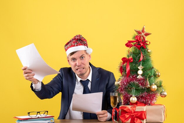 Vista frontal del hombre enojado con gorro de Papá Noel sentado en la mesa cerca del árbol de Navidad y presenta en el espacio de copia de pared amarilla