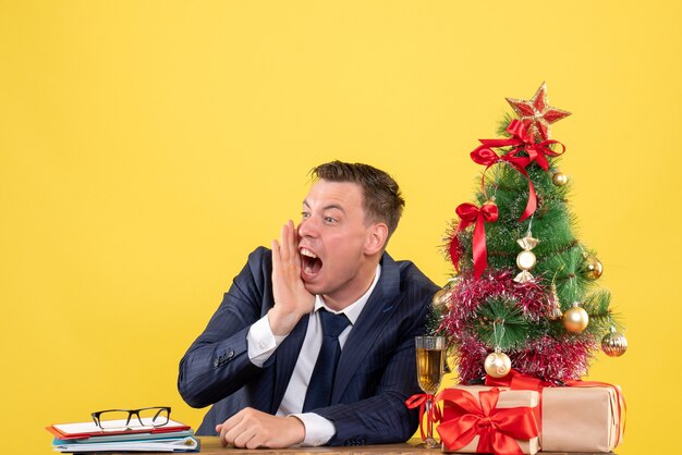 Vista frontal del hombre enojado gritando mientras está sentado en la mesa cerca del árbol de Navidad y presenta en la pared amarilla