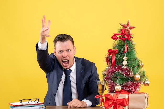 Vista frontal hombre enojado gritando mientras está sentado en la mesa cerca del árbol de Navidad y regalos sobre fondo amarillo