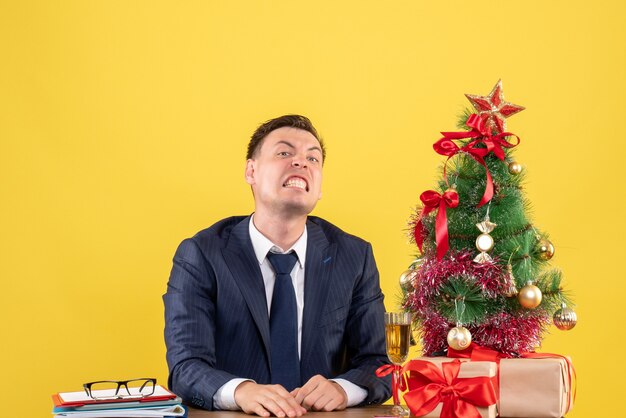 Vista frontal del hombre enojado sentado en la mesa cerca del árbol de Navidad y presenta en el lugar de copia de pared amarilla